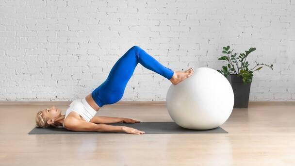 Mulher demonstrando o exercício elevação pélvica com bola. Está deitada no chão, elevando o quadril, enquanto apoia os pés em uma bola de pilates.